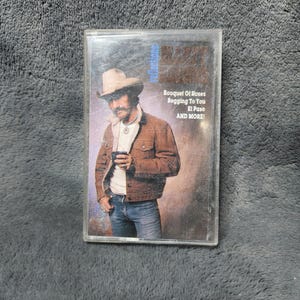 Op de afbeelding: Een vintage cassettebandje met een countrymuziekalbumhoes. De afbeelding toont een man met een cowboyhoed en jas. De tekst op de hoes luidt "Bouquet Of Roses, Begging To You, El Paso, AND MORE!"