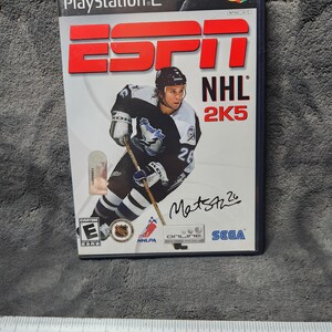 May include: A PlayStation 2 video game case for ESPN NHL 2K5. The cover features a hockey player in a black and white uniform with the number 26. The game's title and the ESPN logo are in red.