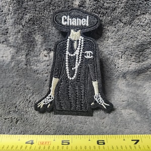 May include: An embroidered patch featuring a figure in a black dress, a wide-brimmed hat with the text Chanel, and long pearl necklaces. The patch is positioned above a yellow measuring tape showing a width of approximately 3.25 inches.
