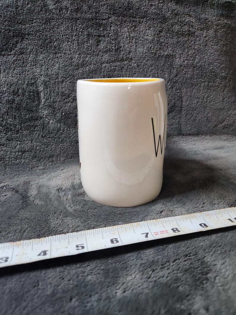 Puede incluir: Una taza de cer&aacute;mica blanca con interior amarillo. La palabra "Wine" est&aacute; escrita en escritura negra en el lateral. Una cinta m&eacute;trica est&aacute; en primer plano.