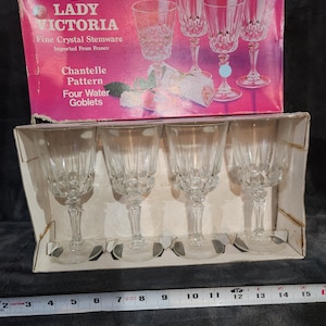 May include: A set of four clear crystal water goblets in a box. The box is pink and has the words "LADY VICTORIA" and "Chantelle Pattern Four Water Goblets" printed on it. The goblets are made of fine crystal stemware.