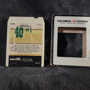 May include: An off-white 8-track tape cartridge labeled "40 #1 Original Country Goldies" with a list of artists. Next to it is an empty Columbia TC8 Stereo 8-track tape cartridge case.