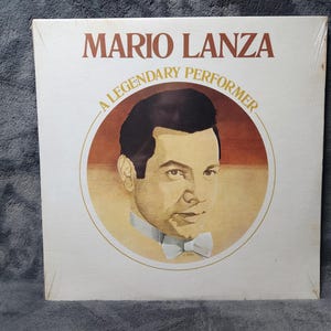 Pode incluir: Um álbum de vinil vintage com Mario Lanza. A capa do álbum apresenta o texto "MARIO LANZA" em letras marrons acima de um retrato do cantor. O retrato está dentro de uma moldura circular com o texto "A LEGENDARY PERFORMER".