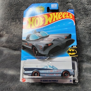 May include: A Hot Wheels die-cast toy car of the TV Series Batmobile, in a blue and black color scheme. The packaging includes the Hot Wheels logo and the text "TV Series Batmobile". The car has red-rimmed wheels and is marked 105/250.