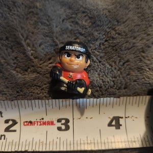 May include: A small, detailed figurine of a hockey player wearing a red jersey and black helmet with the word "SENATORS" on it. The figurine is holding a hockey stick and is approximately 2 inches tall.
