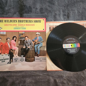 May include: Vintage vinyl record album cover for "The Wilburn Brothers Show" featuring Loretta Lynn, Harold Morrison, and Ernest Tubb. The album cover has a yellow and red border with a photo of the band. A black vinyl record is next to the album.