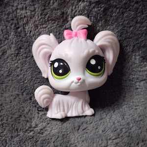 Puede incluir: Figura de perro de juguete rosa claro con grandes ojos negros y blancos, detalles verdes y un lazo rosa. El perro tiene orejas grandes y caídas y una cola rizada. El juguete es de plástico y tiene un acabado liso.