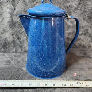 May include: A blue enamel coffee pot with a lid and handle. The pot is approximately 12 inches tall and has white speckles. The spout is on the left side of the pot. The handle is attached to the side of the pot.