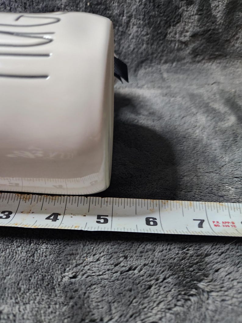 K&ouml;nnte beinhalten: Ein wei&szlig;er rechteckiger Keramikbeh&auml;lter mit einem schwarzen Band. Der Beh&auml;lter wird mit einem Ma&szlig;band gemessen, das eine Breite von etwa 10 cm zeigt.