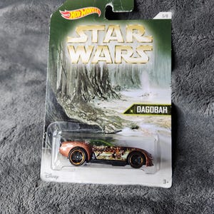 May include: A Hot Wheels Star Wars Dagobah die-cast car in its original packaging. The car is copper-colored with gold wheels and a green top. The packaging features the Star Wars logo and a forest scene.