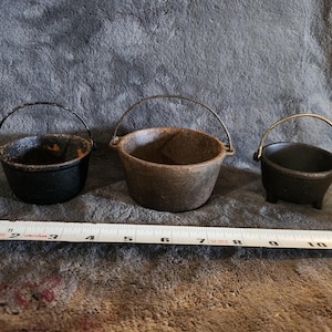 Könnte beinhalten: Drei kleine gusseiserne Kessel mit Metallgriffen. Die Kessel sind schwarz und braun, mit einem Maßband im Vordergrund. Die Kessel haben einen Durchmesser von etwa 7,6 bis 10,2 cm.