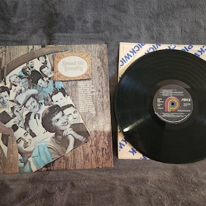 May include: A vinyl record album cover featuring a group of country music singers. The album cover is titled "Grand Ole Country".
