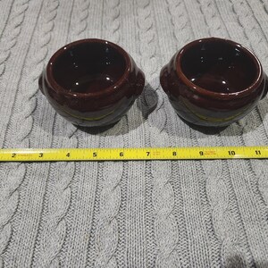 Puede incluir: Dos pequeños cuencos de cerámica marrón con forma redondeada y un borde ligeramente acampanado. Los cuencos están sobre una superficie de punto gris.