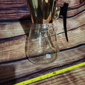 May include: A glass pitcher with a gold-toned finish and a black plastic handle. The pitcher has a gold-coloured lid and a floral design.