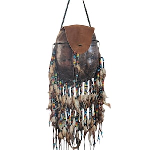 Op de afbeelding: Een handgemaakte, bruinleren tas met een klepsluiting en een lange, gevlochten riem. De tas is versierd met kleurrijke kralen en veren, wat een bohemien esthetiek creëert. De tas is ongeveer 25 cm hoog en 20 cm breed.