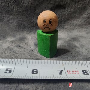 Pode incluir: Uma figura de brinquedo de madeira com uma cabeça redonda e castanha com um rosto triste e sardas, em cima de um corpo geométrico verde. A figura está perto de uma fita métrica, com medidas em centímetros.