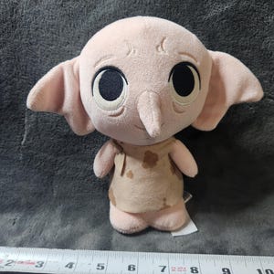 May include: A plush doll of a character with large black eyes, large ears, and a long nose. The doll is primarily pink with a tan outfit. A measuring tape is placed below the doll.