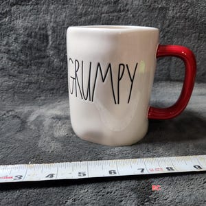 Puede incluir: Taza de cerámica blanca con la palabra "GRUMPY" en letras negras. La taza tiene un asa roja. La taza mide aproximadamente 10 cm de alto.