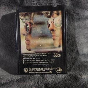 May include: A vintage black 8-track tape of George Harrison's album "Thirty-Three & 1/3". The cover features a side profile of a man wearing sunglasses. The tracklist includes songs like "Woman Don't You Cry For Me" and "Dear One".