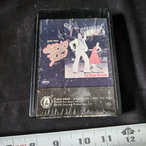 Op de afbeelding: Een zwarte cassettebanddoos met een afbeelding van een dansend stel. De doos heeft de tekst "Music from Saturday Night Fever" en "Performed by The Music Machine".