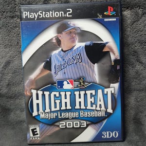 Puede incluir: Una caja de videojuego de PlayStation 2 para "High Heat Major League Baseball 2003". La portada presenta a un jugador de béisbol con uniforme de Arizona. El título del juego está en letras blancas grandes sobre un fondo azul. El juego está clasificado "E" para Todos.