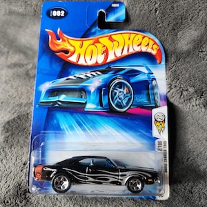 Puede incluir: Un coche de juguete Hot Wheels Dodge Charger 1969 en su embalaje original. El coche negro tiene gráficos de llamas plateadas y detalles en naranja. El embalaje es azul con el logotipo de Hot Wheels.