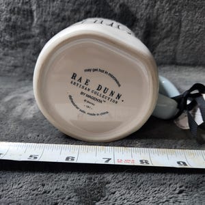 K&ouml;nnte beinhalten: Wei&szlig;er Keramikbecher mit hellblauem Henkel. Auf der Unterseite des Bechers steht der Text "RAE DUNN ARTISAN COLLECTION BY MAGENTA" und "may get hot in microwave". Ein schwarz-wei&szlig;es Band ist am Henkel befestigt.