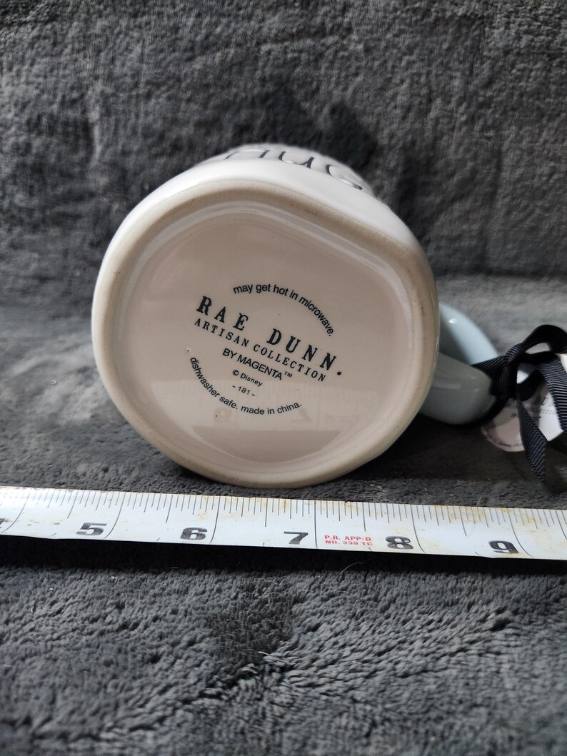 K&ouml;nnte beinhalten: Wei&szlig;er Keramikbecher mit hellblauem Henkel. Auf der Unterseite des Bechers steht der Text "RAE DUNN ARTISAN COLLECTION BY MAGENTA" und "may get hot in microwave". Ein schwarz-wei&szlig;es Band ist am Henkel befestigt.