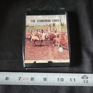 Peut inclure: Une cassette audio vintage avec un boîtier noir et une image colorée d'un cow-boy à cheval dans un champ avec du bétail. Le texte "THE STONEMAN FAMILY" est imprimé sur la face avant de la cassette.