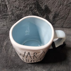 K&ouml;nnte beinhalten: Wei&szlig;er Keramikbecher mit hellblauem Innenraum und Henkel. Auf dem Becher steht in schwarzer Schrift "WARM HUGS". Der Becher steht auf einer strukturierten grauen Oberfl&auml;che.