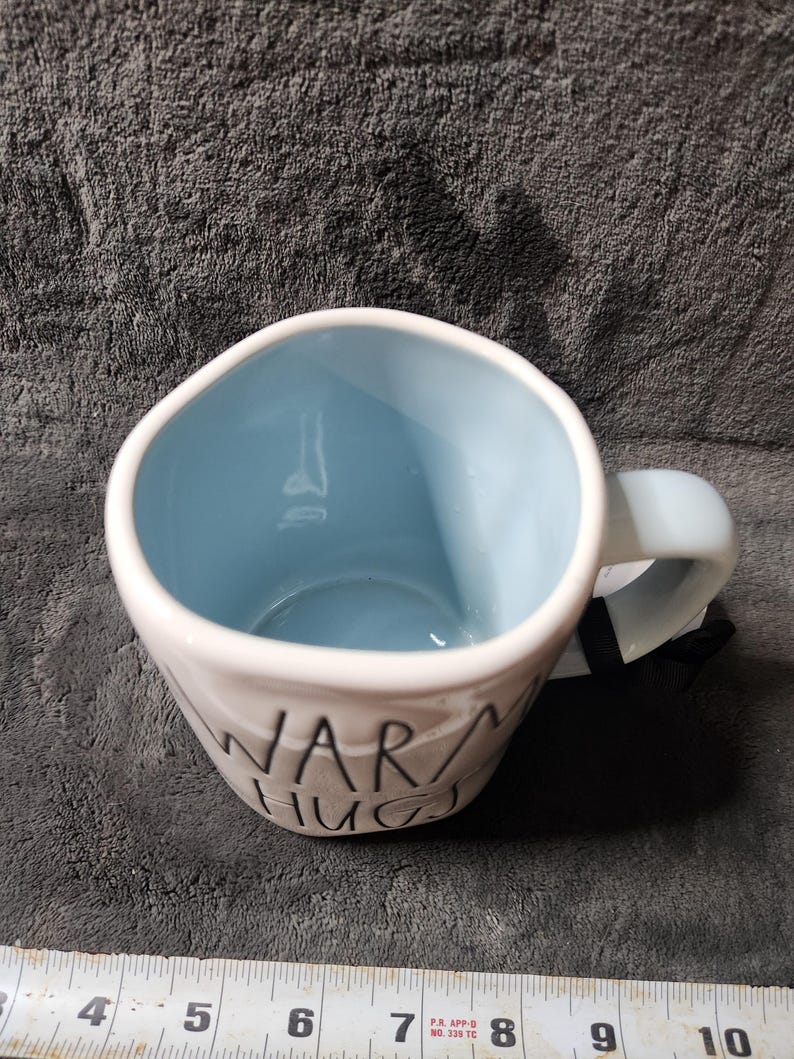 K&ouml;nnte beinhalten: Wei&szlig;er Keramikbecher mit hellblauem Innenraum und Henkel. Auf dem Becher steht in schwarzer Schrift "WARM HUGS". Der Becher steht auf einer strukturierten grauen Oberfl&auml;che.