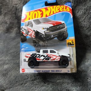 May include: A white and red Hot Wheels die-cast model of a 2019 Chevy Silverado Trail Boss LT truck. The toy truck has black wheels and the word "Borla" printed on the side. The packaging is blue, red, and yellow.