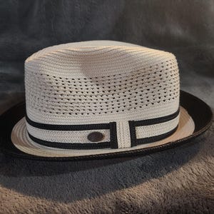 Pode incluir: Um chapéu branco estilo fedora com aba preta e uma faixa preta com um pequeno emblema circular. O chapéu tem uma textura tecida e um topo plano. Um acessório elegante.