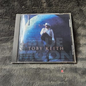 May include: A CD case featuring the album "Blue Moon" by Toby Keith. The cover art depicts a man in a white hat and shirt holding a guitar, set against a blue and dark background. The artist's name is prominently displayed.