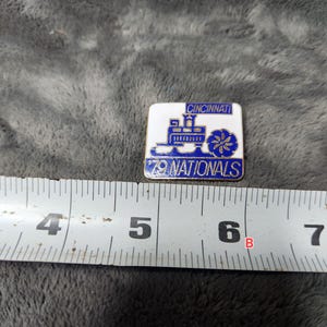 May include: A square pin with a white background and blue accents. The pin features the text "CINCINNATI" at the top and "79 NATIONALS" at the bottom. A blue train and floral design are in the center. The pin measures approximately 1 inch by 1 inch.