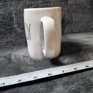 Puede incluir: Taza de cer&aacute;mica blanca con asa curva. La palabra "JOY" est&aacute; impresa en negro en el lateral. Una cinta m&eacute;trica se coloca cerca de la taza.