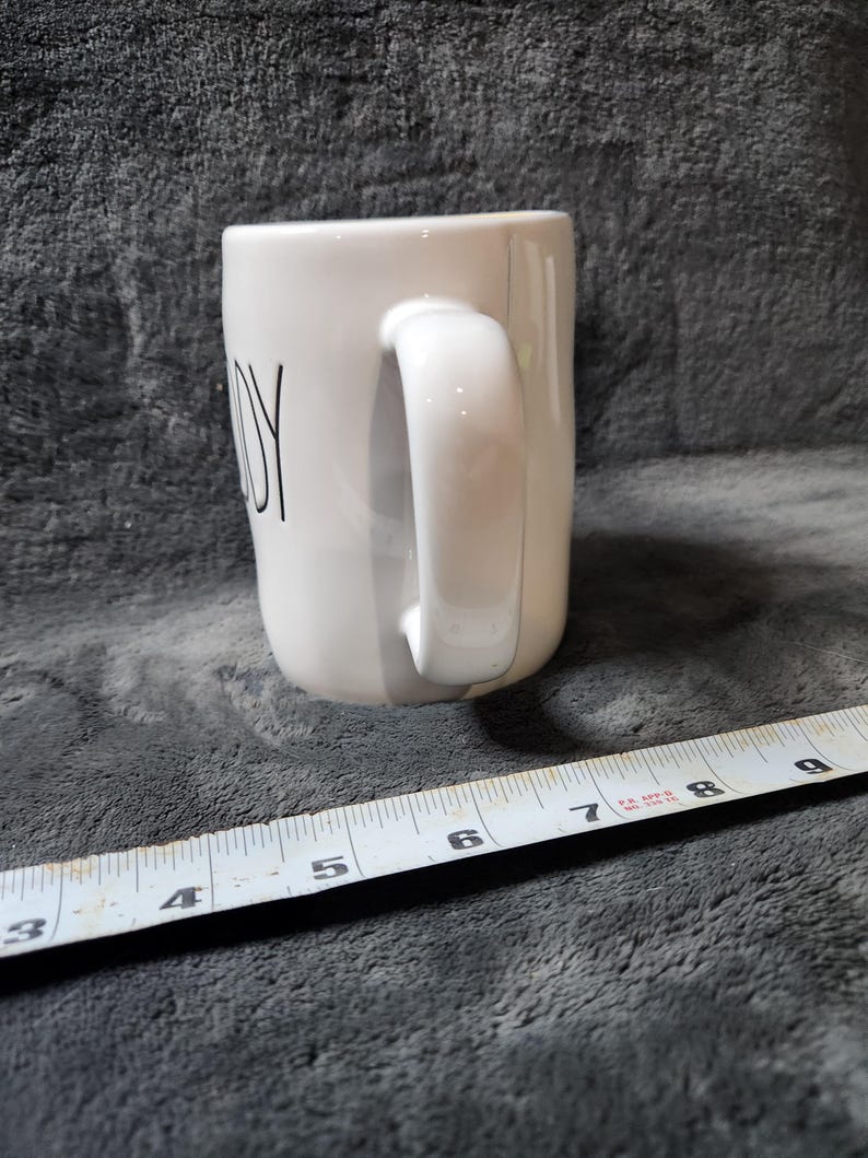 Puede incluir: Taza de cer&aacute;mica blanca con asa curva. La palabra "JOY" est&aacute; impresa en negro en el lateral. Una cinta m&eacute;trica se coloca cerca de la taza.
