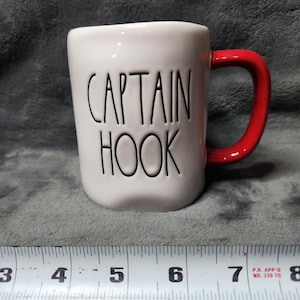 Może przedstawiać: Biały ceramiczny kubek z napisem "CAPTAIN HOOK" w czarnych literach. Kubek ma czerwony uchwyt. Obok kubka znajduje się linijka. Kubek ma około 10 cm wysokości.