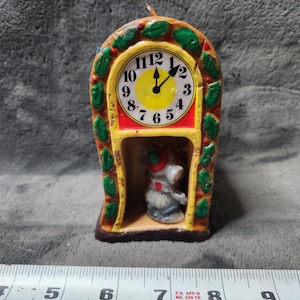 Puede incluir: Una vela vintage con forma de reloj con una esfera amarilla y números negros. El reloj tiene un borde rojo y está decorado con hojas verdes. Una pequeña figura de un ratón está dentro del reloj. La vela mide aproximadamente 15 cm de alto.