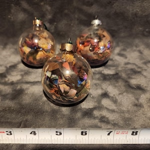 May include: Three clear glass baubles filled with colourful, dried, and pressed leaves and flowers. The baubles are a mix of brown, green, red, and gold.