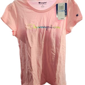 Puede incluir: Camiseta Champion rosa claro con cuello redondo y mangas cortas. El logo de Champion está impreso en el pecho en degradado arcoíris. Pequeño logo de Champion en la manga izquierda. Etiqueta de precio adjunta.