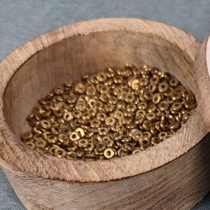 May include: A round wooden container with a light-colored lid featuring a decorative pattern, filled with small, metallic gold-toned circular beads. Several identical gold beads are scattered outside the container on the surface.