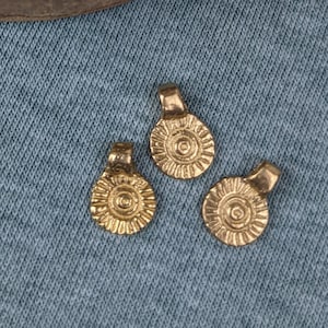 May include: A wooden bowl filled with gold-colored metal charms with a circular design. Three charms are displayed on a blue fabric surface.