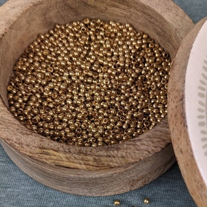 以下が含まれることがあります： 装飾的なデザインが施された白い蓋が付いた木製の箱。箱は小さな金色のビーズでいっぱい。いくつかのビーズが青い表面に散らばっている。