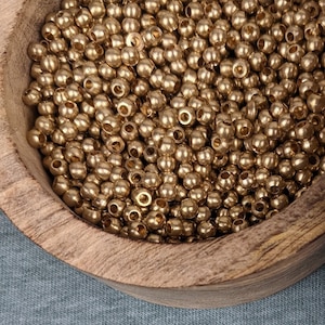 Puede incluir: Una caja de madera con tapa que contiene una gran cantidad de pequeñas cuentas doradas. La tapa tiene un patrón decorativo en tonos rosa y gris. Algunas cuentas están esparcidas sobre una superficie de tela azul.