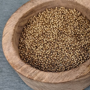 Op de afbeelding: Een houten kom gevuld met kleine, gouden kralen, met wat kralen verspreid op een blauwgroen oppervlak. De kom is open en onthult de inhoud. Het deksel, met een decoratief ontwerp, is gedeeltelijk zichtbaar.