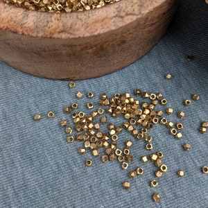 Könnte beinhalten: Eine Holzkiste gefüllt mit goldfarbenen Metallperlen. Die Perlen sind klein und haben eine facettierte, diamantartige Form. Einige Perlen sind auf einer blauen Stoffoberfläche verstreut.