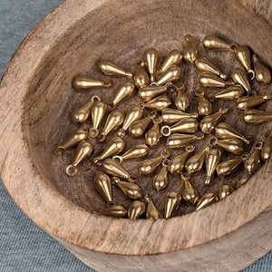 May include: A wooden box with a lid, filled with gold teardrop-shaped charms. The lid has a white background with a grey floral design. Three gold teardrop charms are on a grey surface.