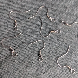 Pode incluir: Um conjunto de seis ganchos para brincos em metal prateado, cada um com um fio curvo, uma pequena espiral decorativa e um laço circular na parte inferior para prender componentes de joalharia, dispostos de forma dispersa.