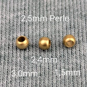 May include: Three gold-colored metal beads of different sizes. The beads are 2.5mm, 2.4mm, and 1.5mm in diameter. The beads are on a gray textured surface.